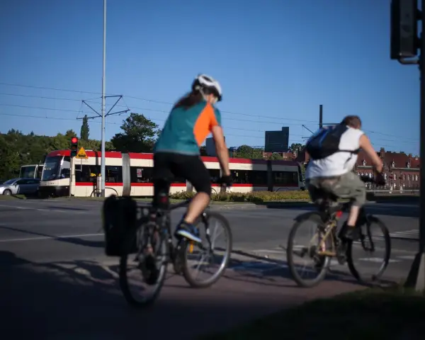 Rowerzyści jadący wzdłuż torów tramwajowych, w tle czerwono-biały tramwaj. Trasa rowerowa wiodąca w kierunku Westerplatte.