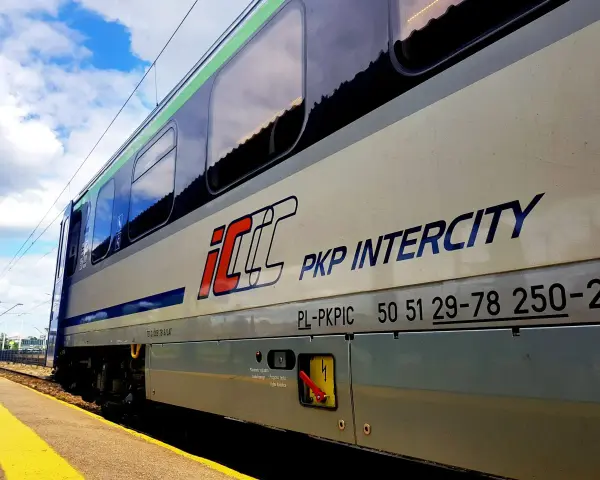 Pociąg PKP Intercity z napisem "Warszawa Poznań" na tle błękitnego nieba z chmurami.