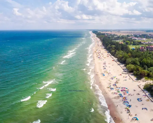 Słoneczny dzień na plaży w Sarbinowie nad morzem. Tłumy turystów odpoczywają na piasku, kąpią się w morzu i spacerują wzdłuż brzegu.