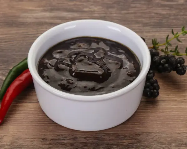 Gęsty, ciemny sos pieprzowy w białej miseczce, obok czerwona i zielona papryczka chili oraz gałązka jeżyn.