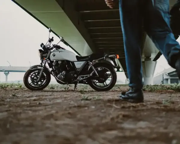 Biały motocykl klasyczny, kategoria A2, pod mostem. Obok przechodzi motocyklista z kaskiem.