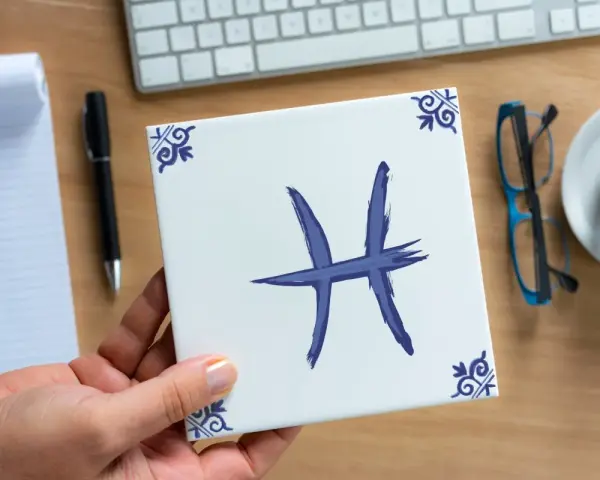 Dłoń trzyma kwadratową płytkę z symbolem ryby, znakiem zodiaku. Na biurku leżą notatnik, długopis, okulary i kawa.