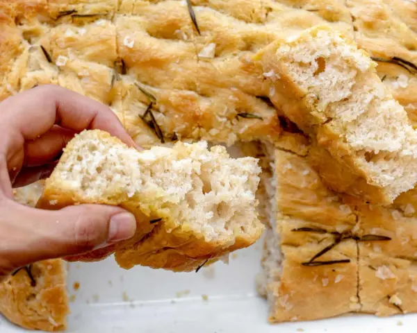 Mano rompiendo un trozo de focaccia casera con romero y sal gruesa. ¡Una delicia de esta focaccia receta!