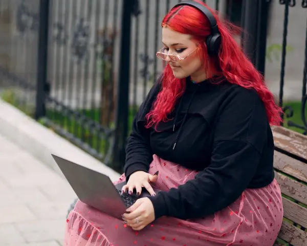 Młoda babista z ognistymi włosami i słuchawkami na uszach pracuje na laptopie, siedząc na ławce w parku.