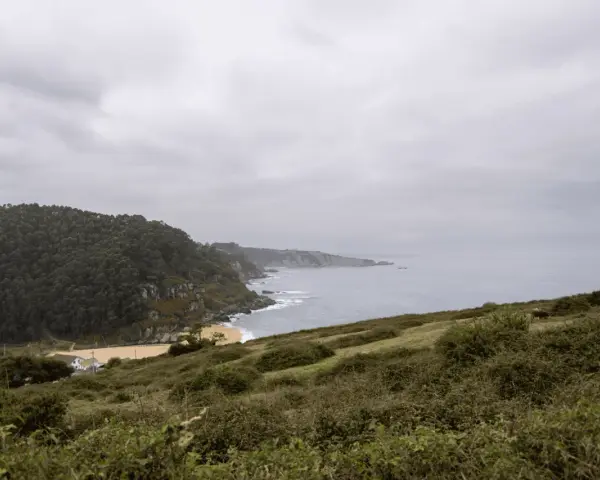 Paisaje costero con acantilados verdes y mar gris. Ideal para senderismo en Villaviciosa, con una playa dorada y una casa blanca.