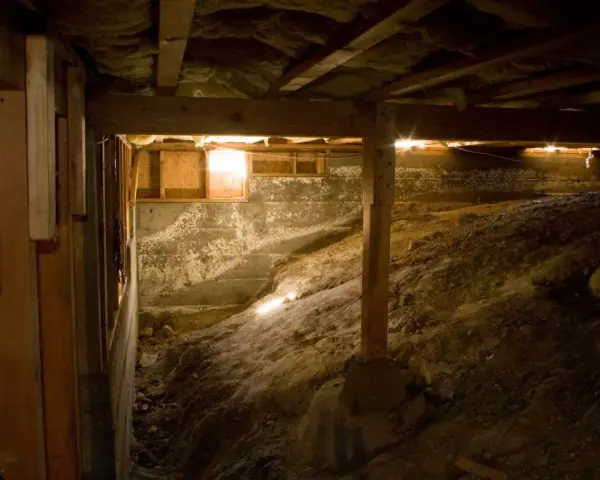 Underneath a house, a crawl space with a dirt floor and concrete walls. A vapor barrier is being installed to protect against moisture.
