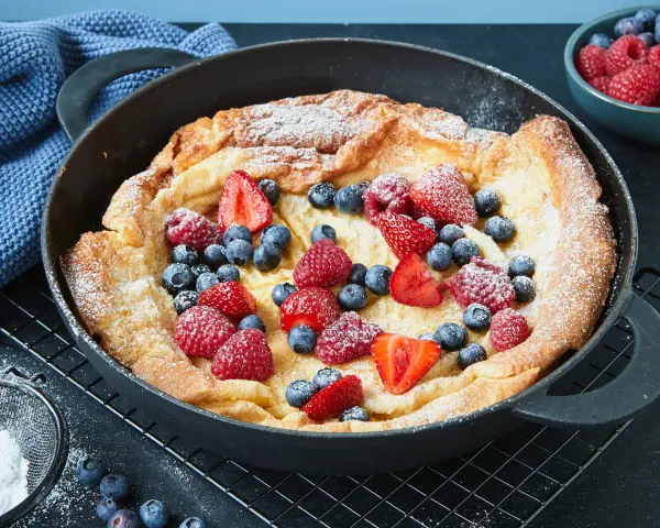 Ein goldbrauner Dutch Baby mit Puderzucker und frischen Beeren, serviert in einer gusseisernen Pfanne.