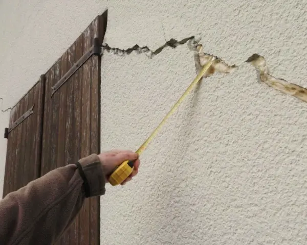 Main d'une personne mesurant des fissures sur un mur extérieur. Quand s'inquiéter des fissures maison ?