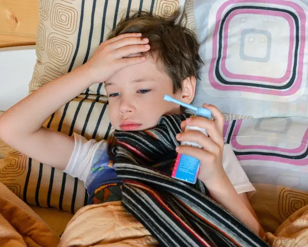Chory chłopiec leży w łóżku z ręką na czole i trzyma inhalator.
