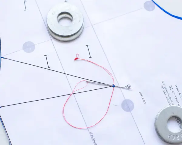 Sur un patron de couture, une aiguille rouge traverse le papier, retenue par des rondelles métalliques.