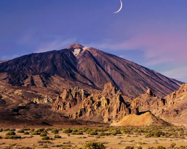 Krajobraz w parku narodowym Teide z księżycem w kształcie sierpa na fioletowym niebie. Wulkan góruje nad skalistym terenem.