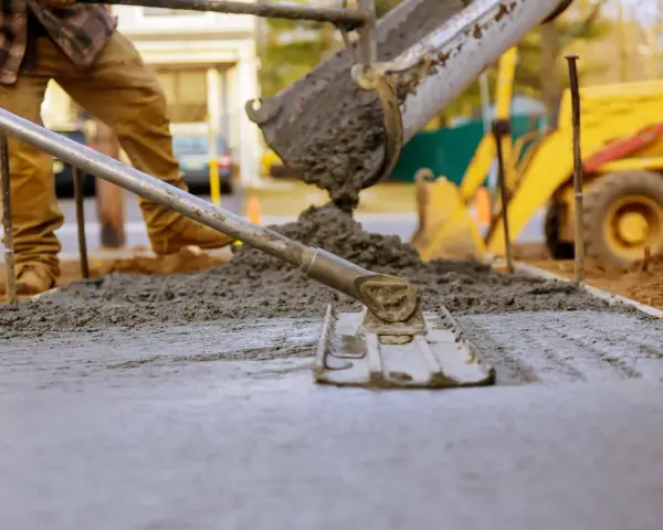 Pracownik wylewa beton, który będzie schnął. W tle koparka i budowa.