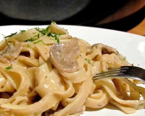 Tagiatelle z grzybami w kremowym sosie, posypane świeżym koperkiem. Widok z bliska na talerz pełen pysznego makaronu.