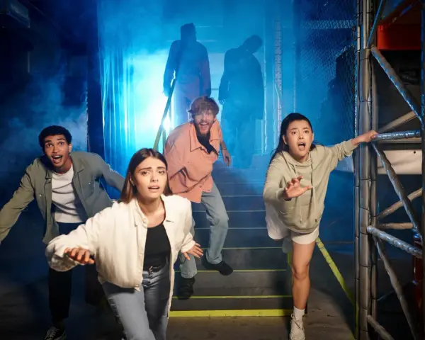 Grupa przerażonych ludzi ucieka przed postaciami w dymie. To wygląda jak prawdziwy escape room, gdzie trzeba rozwiązać zagadki, by przeżyć.