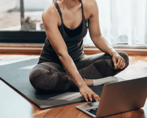 Kobieta w stroju sportowym siedzi na macie, korzystając z laptopa. Czy można ćwiczyć codziennie? Tak, jeśli masz dostęp do inspiracji online!