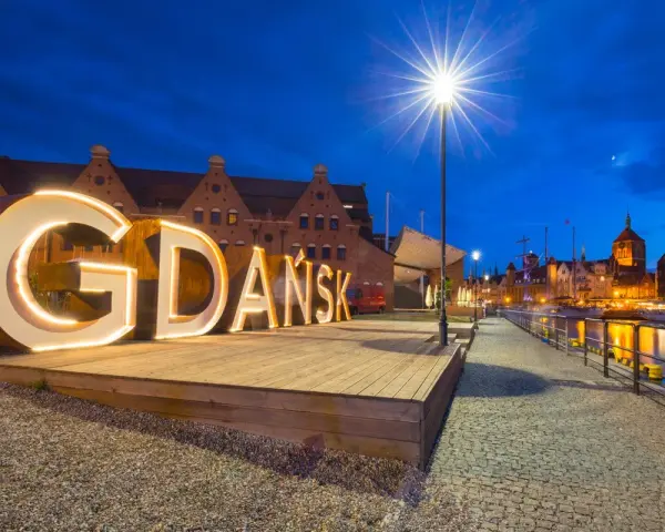 Podświetlany napis "GDAŃSK" na tle nocnej panoramy miasta, zapraszający do odkrywania atrakcji Trójmiasta.
