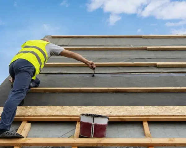 Pracownik w kamizelce zakłada papę na dach z desek, uszczelniając połączenia.