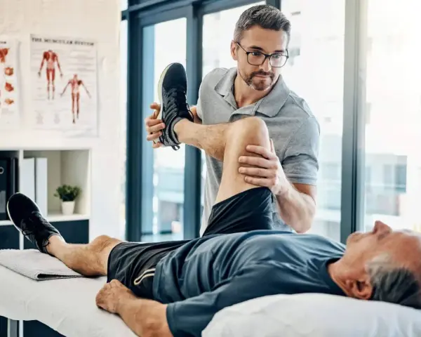 Ein Physiotherapeut dehnt das Bein eines Patienten. Der Patient liegt auf einer Liege, während der Therapeut sein Knie beugt. Wann Physiotherapie? Hier wird geholfen.