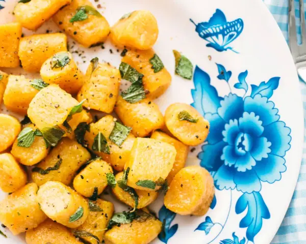 Gnocchi di zucca dorati, conditi con erbe aromatiche e pepe nero, su un piatto decorato con fiori blu.