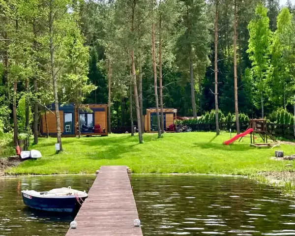 Drewniany pomost nad jeziorem Bardze, z łódką przycumowaną do brzegu. W tle domki letniskowe i plac zabaw.