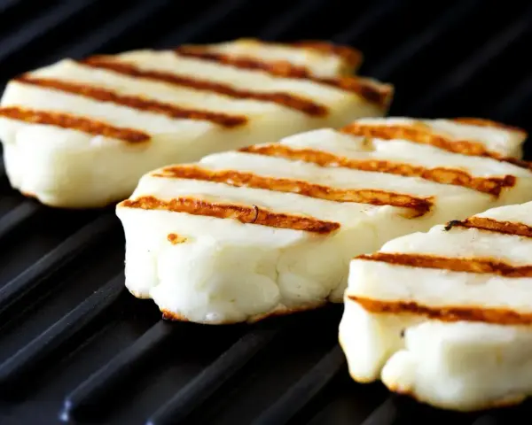 Trzy kawałki halloumi z grilla, z apetycznymi paskami, leżą na czarnej grillowej płycie.
