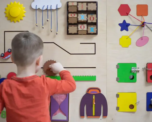 Chłopiec bawi się na tablicy edukacyjnej, rozwijając umiejętności manualne zgodnie z metodą Montessori. Widoczne są kolorowe kształty, zębatki i zamek.