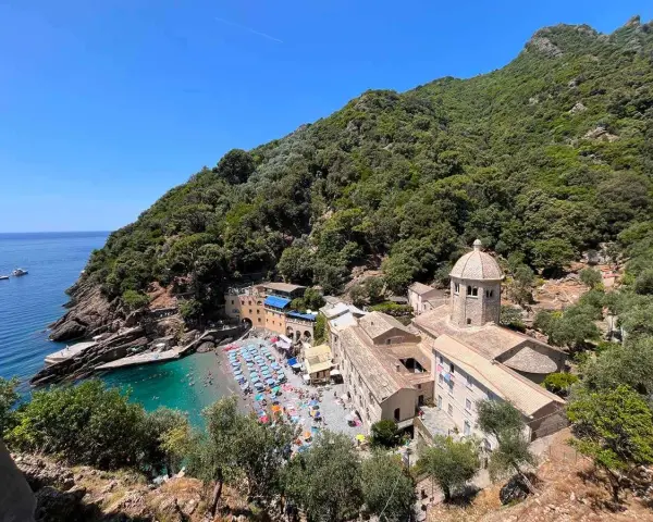 Baia con spiaggia affollata e un antico monastero, un luogo ideale per i cammini in Liguria, con mare azzurro e vegetazione rigogliosa.