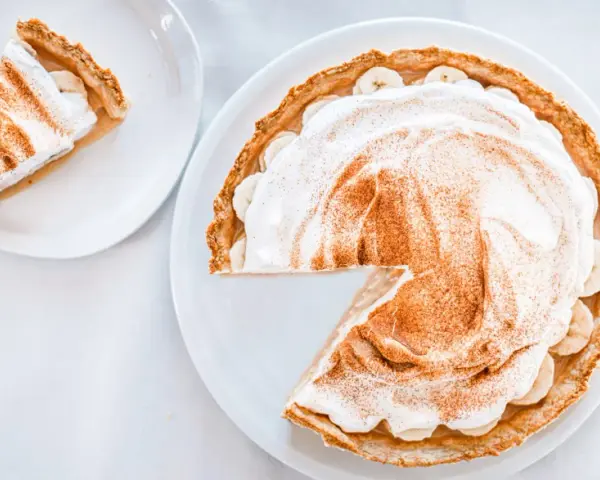 Une part de tarte banoffee traditionnel, garnie de crème fouettée et de cannelle, sur une assiette blanche.