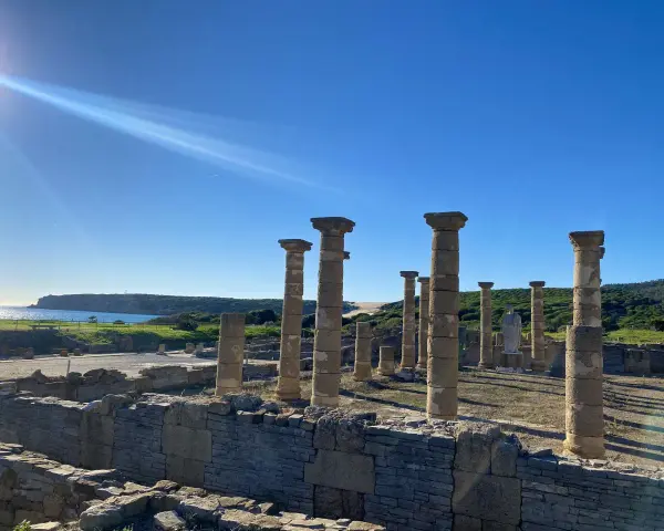 Sunlight streams over ancient roman ruins in Spain, illuminating weathered stone columns and walls against a bright blue sky and distant coastline.