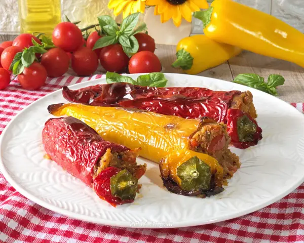 Peperoni cornetti ripieni, rossi e gialli, cotti al forno, su un piatto bianco con pomodorini e basilico.