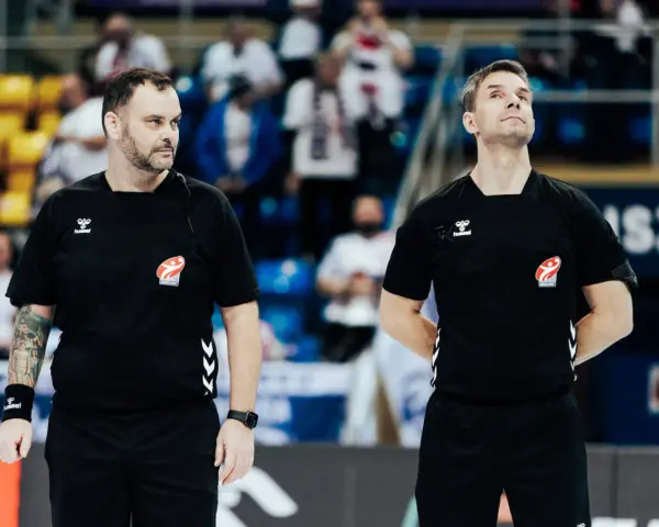 Dwóch sędziów w czarnych koszulkach z logo Hummel i emblematem związku sportowego, gotowych do rozpoczęcia meczu ligi piłki ręcznej.