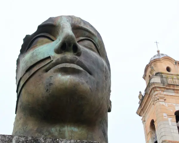Brązowa, monumentalna rzeźba głowy z widocznym fragmentem dzwonnicy kościelnej w tle.