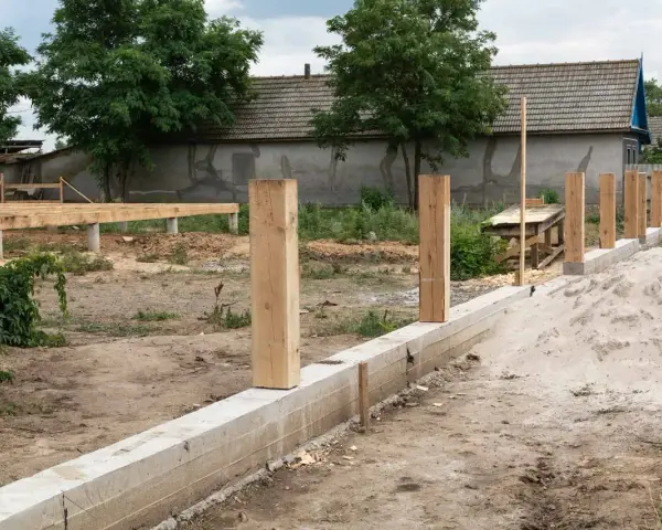 Solidny fundament pod ogrodzenie z drewnianymi słupkami, w tle budowa domu.