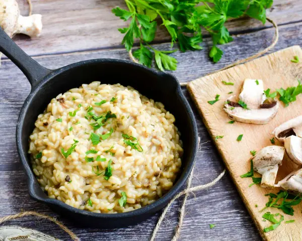 Kremowe risotto z grzybami w czarnej patelni, ozdobione natką pietruszki, obok świeże grzyby na desce.