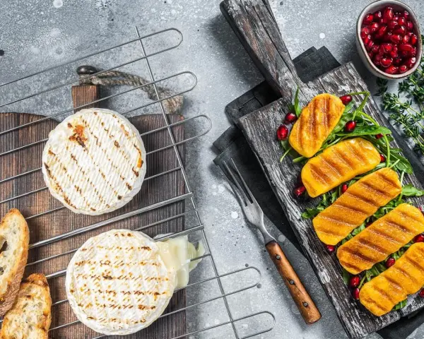 Dwa okrągłe serki na grilla, jeden lekko roztopiony, obok pieczywo. Na desce grillowane kawałki dyni z granatem i rukolą.
