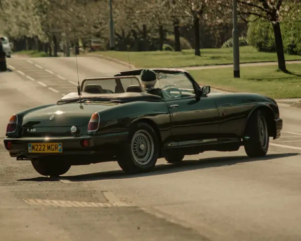 Auto d'epoca verde scuro in movimento, con conducente con cappello. La targa suggerisce che siamo in Inghilterra, dove si guida a destra.
