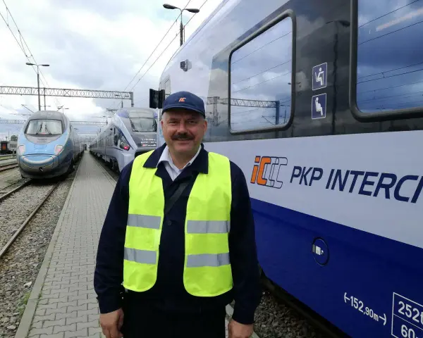 Uśmiechnięty rewident taboru kolejowego w kamizelce odblaskowej stoi obok pociągu PKP Intercity.