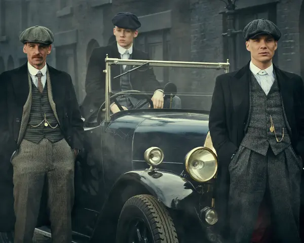 Tre uomini in abiti eleganti, stile Peaky Blinders, posano accanto a un'auto d'epoca.