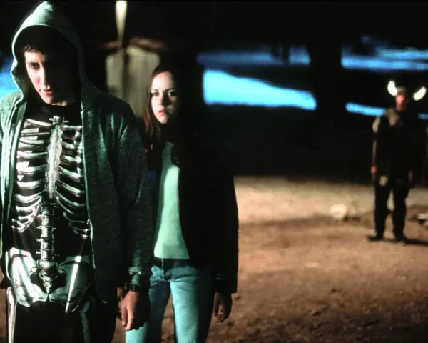 Donnie Darko w bluzie z kapturem i koszulce z nadrukiem szkieletu, obok dziewczyny. W tle dwie postacie w kostiumach.