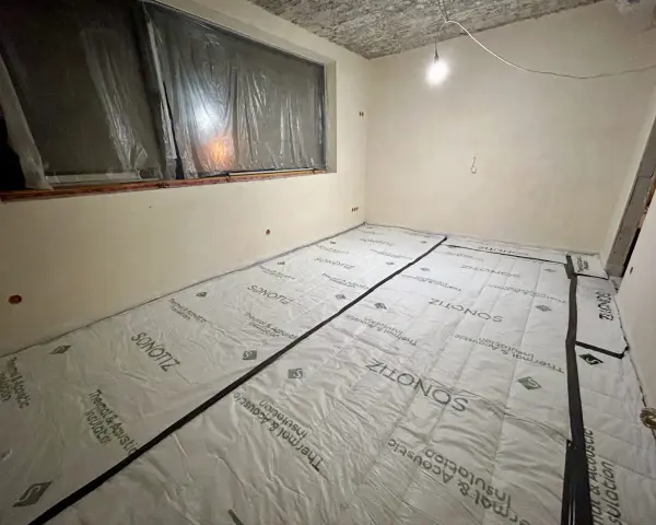 W trakcie prac remontowych w bloku, podłoga pokryta jest materiałem do wyciszenia. Widać okno zabezpieczone folią i sufit z widocznymi instalacjami.