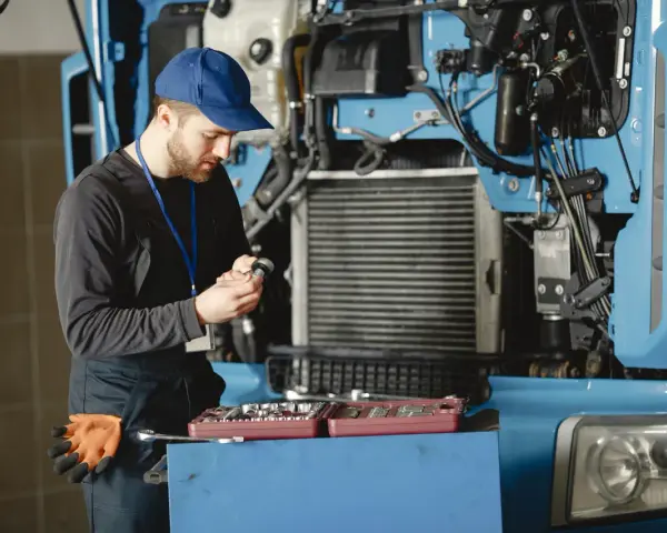 Mechanik w niebieskiej czapce i kombinezonie naprawia ciężarówkę, trzymając narzędzie.