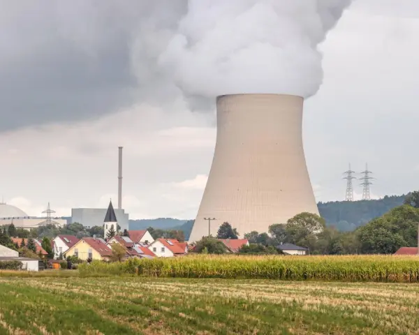 Dampf steigt aus einem Kühlturm eines Atomkraftwerks auf, daneben ein Dorf und ein Maisfeld.