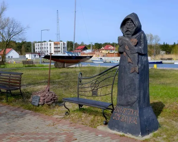 Drewniany pomnik rybaka trzymającego rybę, z napisem "Ławeczka Rybaka". W tle port z łodziami i budynkami.