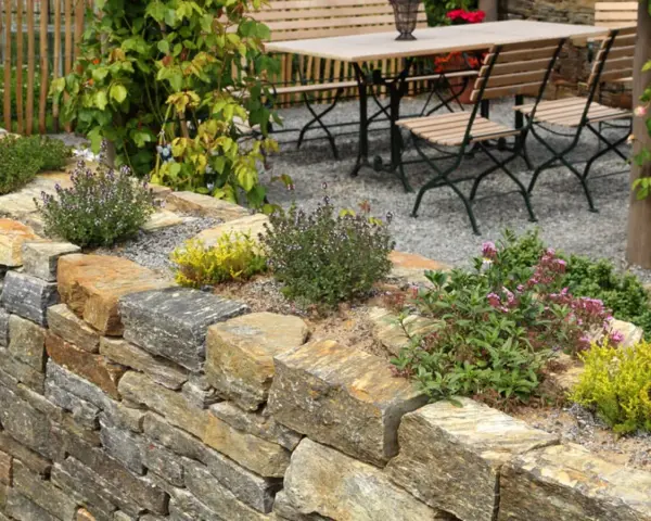 Natürliche Steinmauer mit Kräutern und Blumen, die schöne Ideen für den Garten bietet. Eine Sitzgruppe lädt zum Verweilen ein.