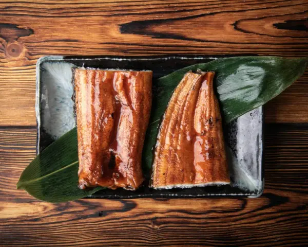 Dwa kawałki glazurowanego węgorza do sushi, podane na liściu na ciemnej zastawie, na drewnianym tle.
