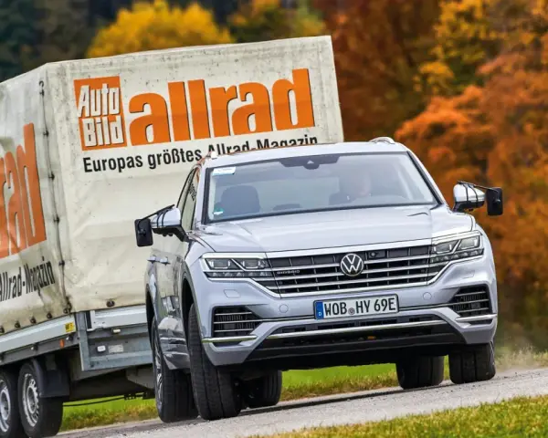 Srebrny Volkswagen Touareg holuje przyczepę z logo "Auto Bild allrad", jadąc po krętej drodze wśród jesiennych drzew.