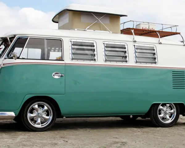 En turkos och vit volkswagen folkabuss med taklucka och takräcke står på en sandstrand.