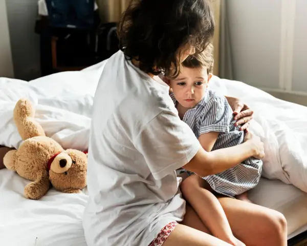 Mutter tröstet ihr Kind, das Anzeichen einer Panikattacke bei Kindern zeigt. Ein Teddybär liegt daneben.