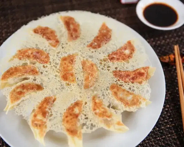 Chrupiące pierożki gyoza ułożone na talerzu, z chrupiącą, złotą panierką. Obok miseczka sosu sojowego i pałeczki.