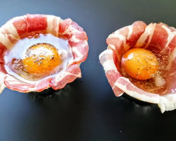 Due uova crude con tuorlo brillante, avvolte in strisce di bacon croccante, pronte per essere cotte.